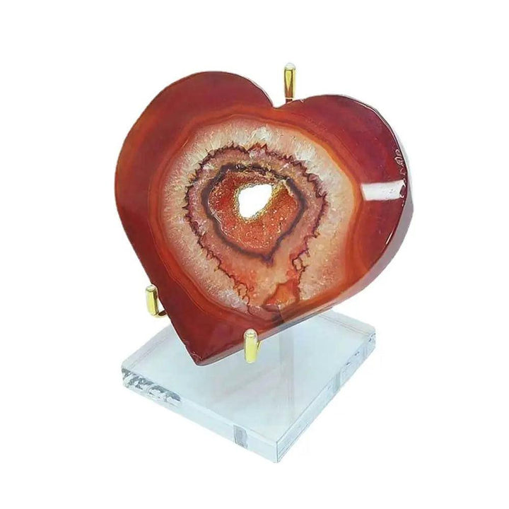 Heart-shaped red agate geode with white and orange bands, displayed on a gold metal and clear acrylic stand.