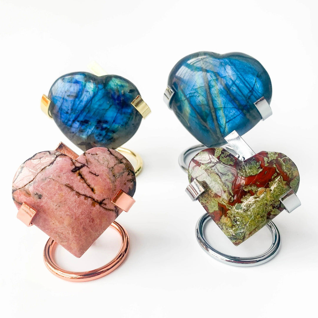 Four heart-shaped crystal spheres: two blue labradorite on gold/silver stands, one pink rhodonite on rose gold, and one green dragon's blood jasper on silver.