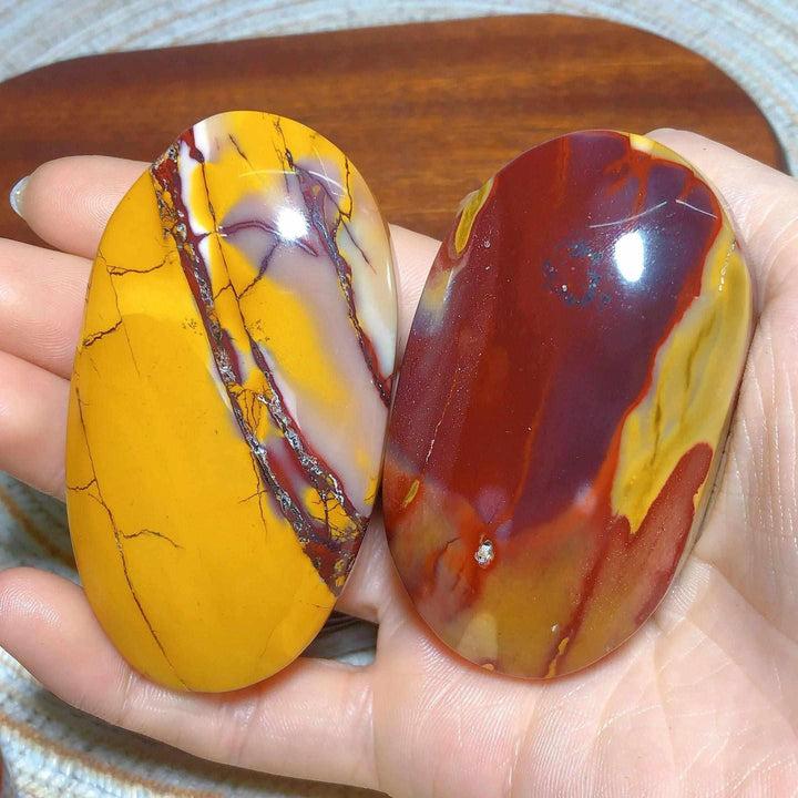 Two smooth, polished Mookaite Jasper palm stones featuring vibrant swirls of red, mustard yellow, and cream for grounding.