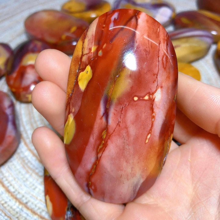 Smooth Mookaite Jasper palm stone in hand, featuring vibrant swirls of red, yellow, cream, and mauve for grounding energy.
