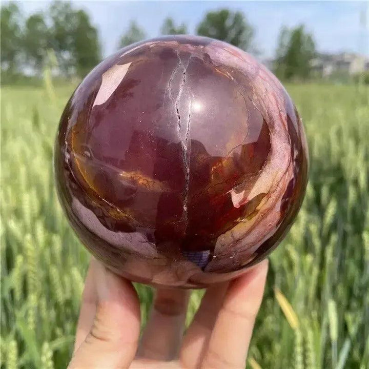 Hand-polished Mookaite Jasper sphere from Crystal Destiny, featuring earthy reds, ivories, and golds, held outdoors.