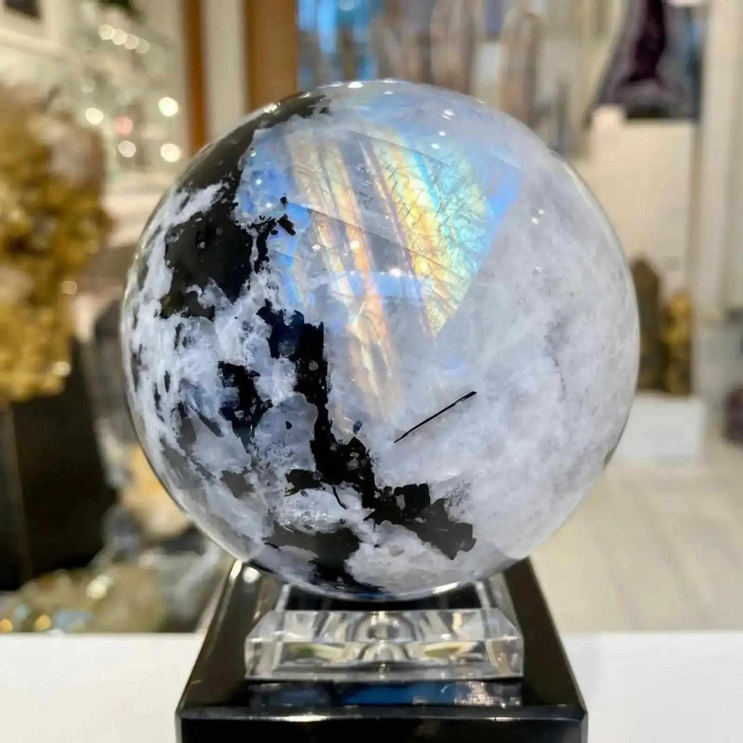 Polished moonstone sphere featuring striking black inclusions and a vibrant, iridescent rainbow flash for crystal healing.