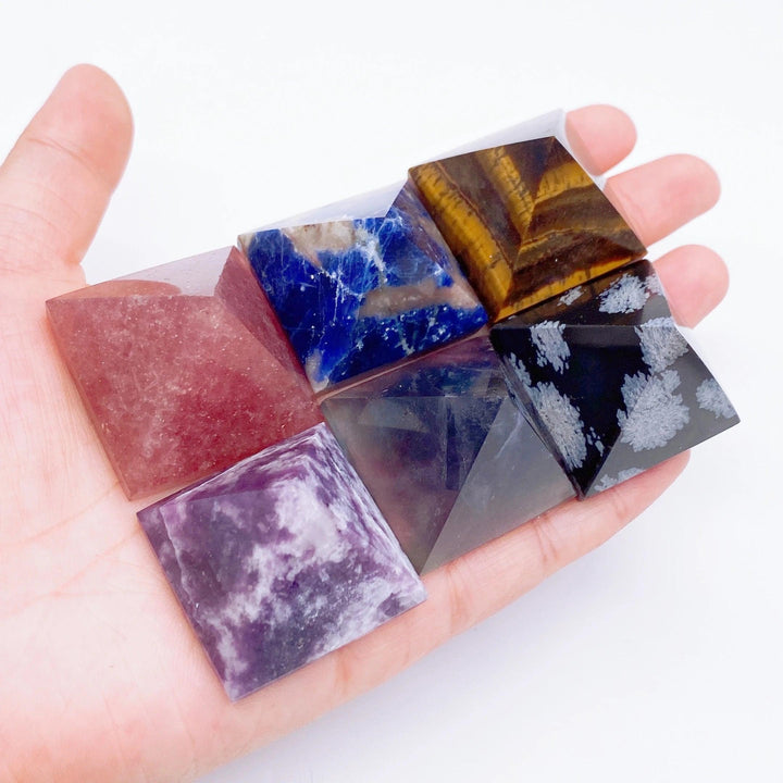Hand holding six natural crystal pyramids: red, blue, tiger eye, snowflake obsidian, dark, and purple for healing.