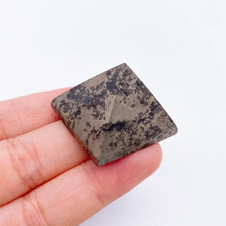 Hand holding a small, dark gray Pyrite crystal pyramid with metallic flecks, ideal for healing and meditation.