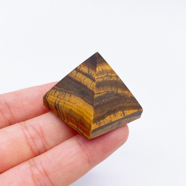 Hand holding a small, golden-brown Tiger Eye crystal pyramid with distinct striped patterns, ideal for healing.