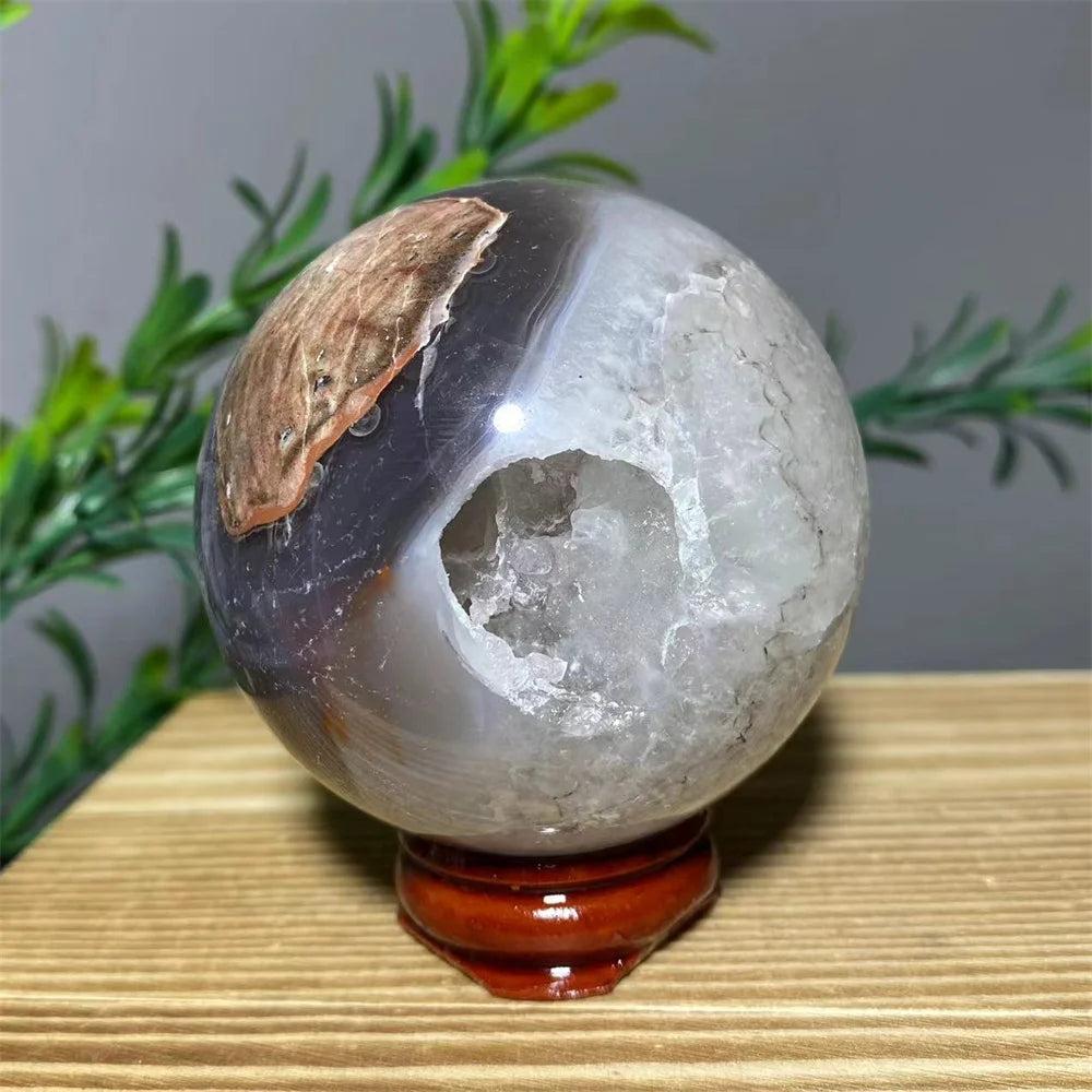 Natural agate crystal sphere with a geode opening, displaying white, gray, and brown tones on a wooden stand.