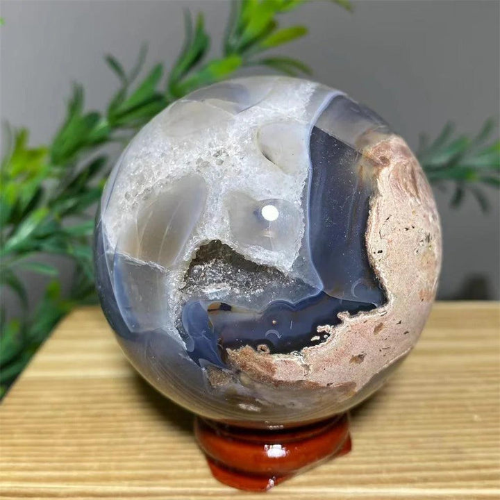 Natural crystal agate sphere with white geode interior, blue-grey bands, and earthy pink tones on a wooden stand.