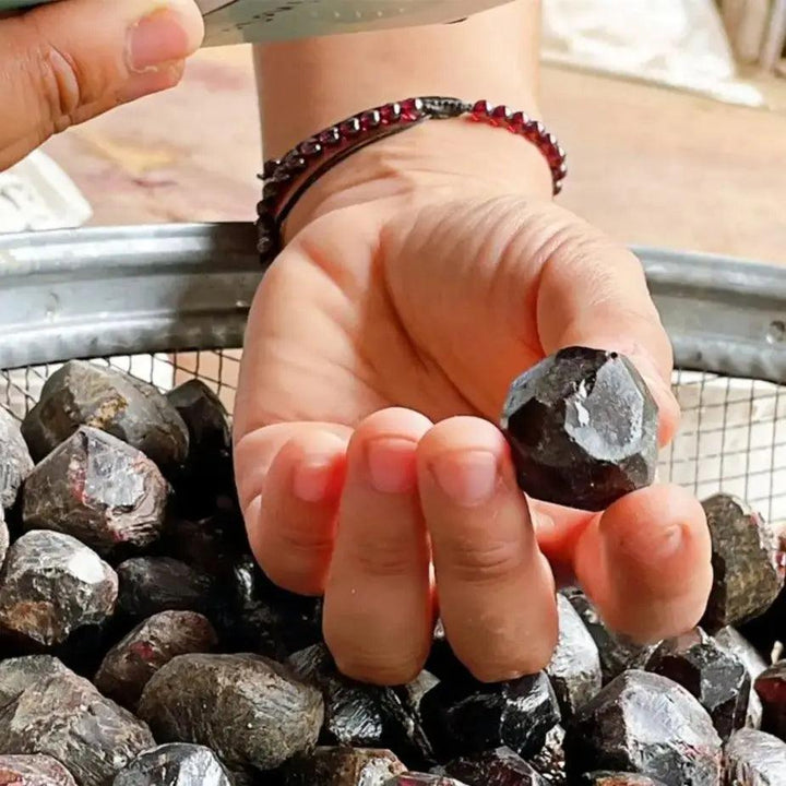 Hand holding a dark red natural rough garnet crystal, surrounded by more raw garnet stones in a metal sieve.