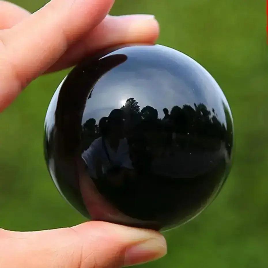 Hand holding a smooth, polished Natural Obsidian Crystal Sphere, reflecting trees and sky.