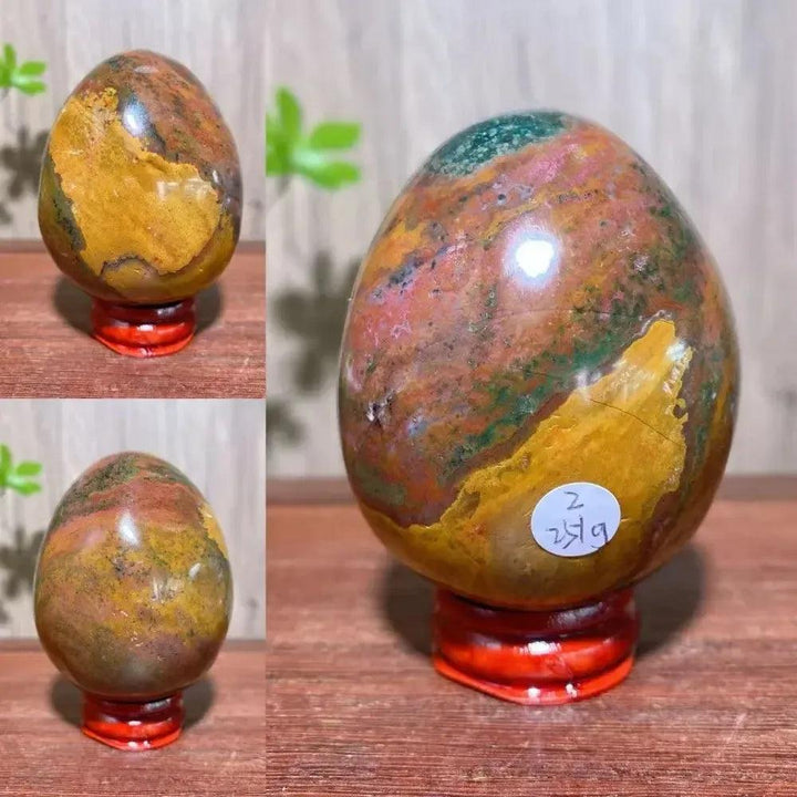 Polished Ocean Jasper crystal egg from Crystal Destiny, displaying greens, pinks, yellows, and browns on a red stand.