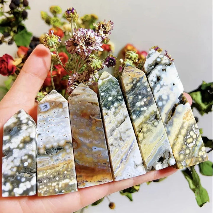 Hand holding six Ocean Jasper crystal towers, 90-100mm, with unique orbicular patterns in sage green, white, coral, and brown.
