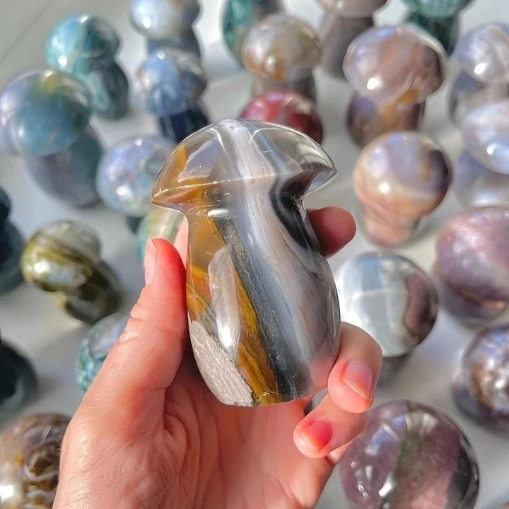 Hand holding a polished Ocean Jasper mushroom carving with sage, cream, and rust swirls, surrounded by more carvings.