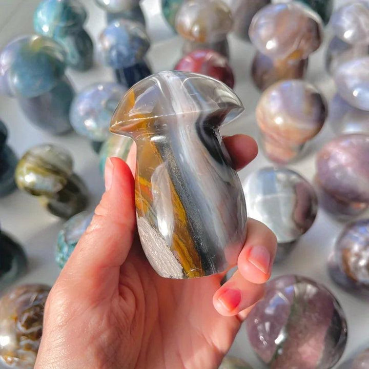 Hand holding a polished Ocean Jasper mushroom carving with sage, cream, and rust swirls, surrounded by similar crystals.