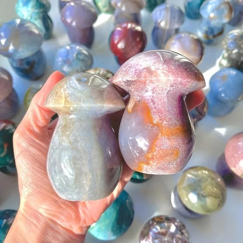 Hand holding two polished Ocean Jasper mushroom carvings, one sage green, one purple and rust, with more in background.
