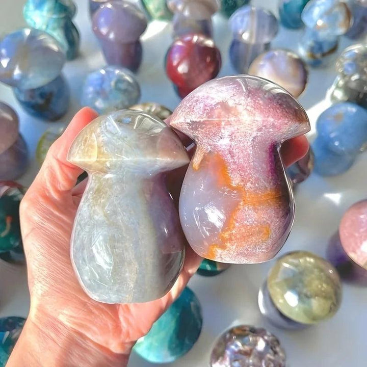 Hand holding two polished Ocean Jasper mushroom carvings, one sage green, one purple and rust, with more in background.