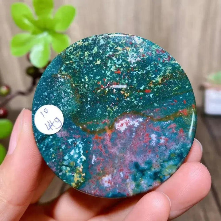 Crystal Destiny's polished Bloodstone round slice, featuring deep greens, reds, and yellows, held in a hand.