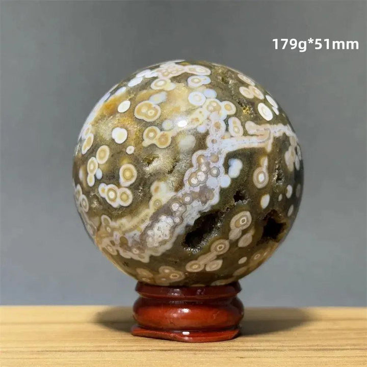 Ocean Jasper sphere from Crystal Destiny, with green, white, and brown orbicular patterns on a red wooden base.