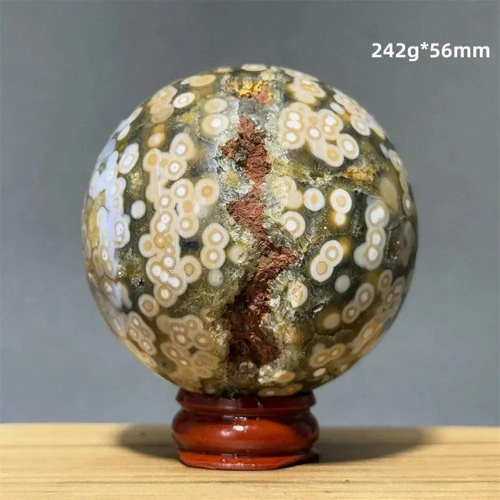 Ocean Jasper sphere from Crystal Destiny, featuring green, white, cream, pink, and red orbicular patterns on a wooden base.
