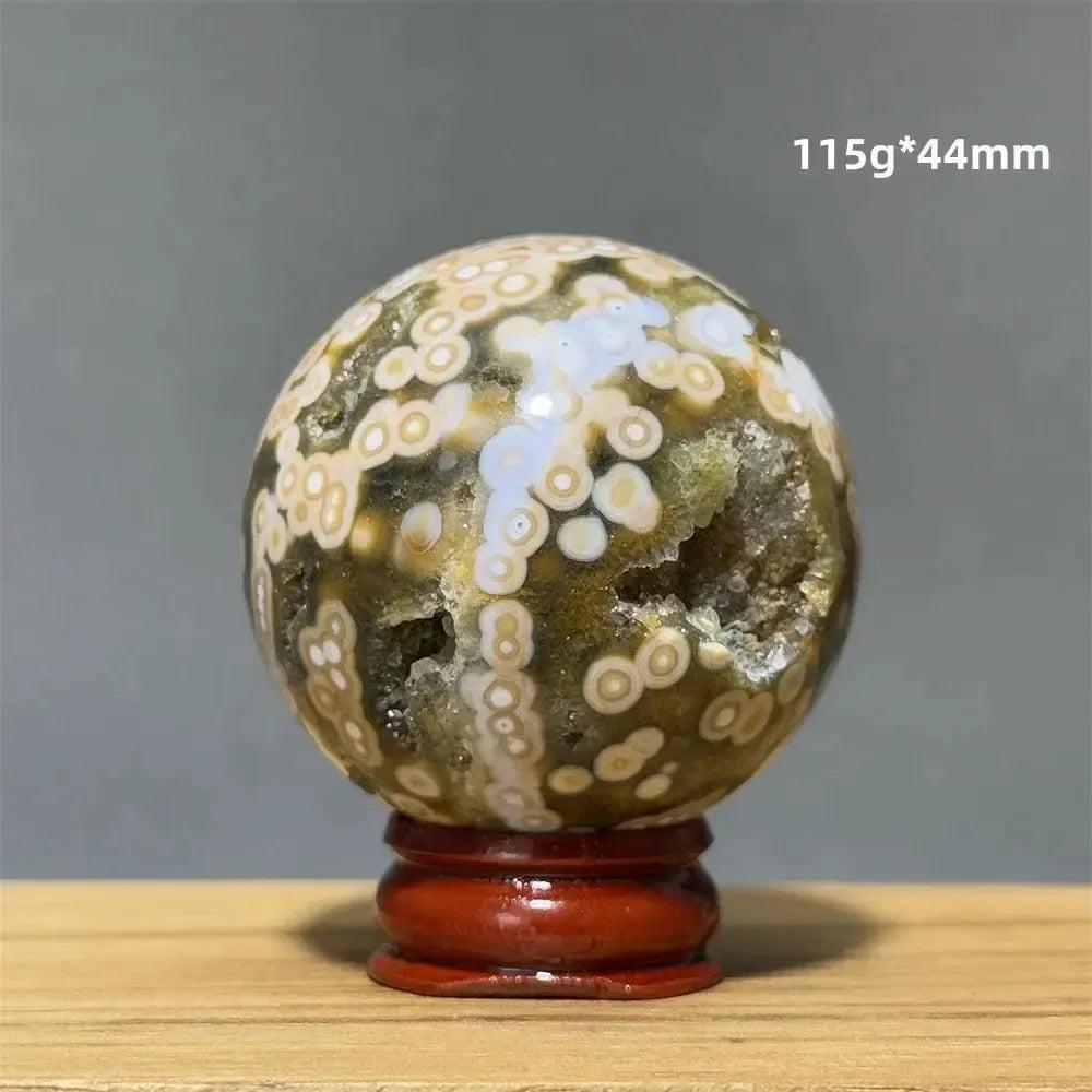 Ocean Jasper sphere from Crystal Destiny, with green, white, and cream orbicular patterns, on a red wooden base.