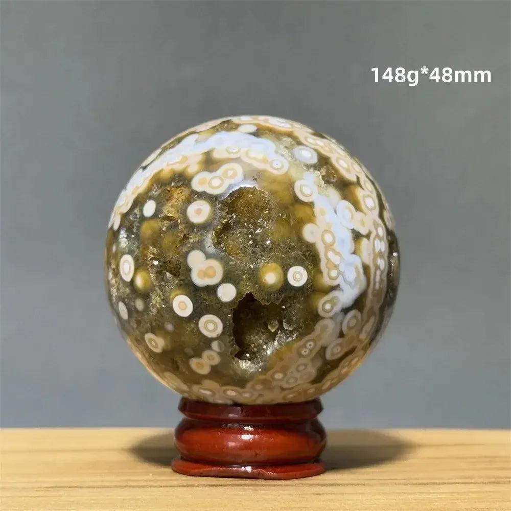 Ocean Jasper sphere with green, white, and brown orbicular patterns on a red wooden base from Crystal Destiny.