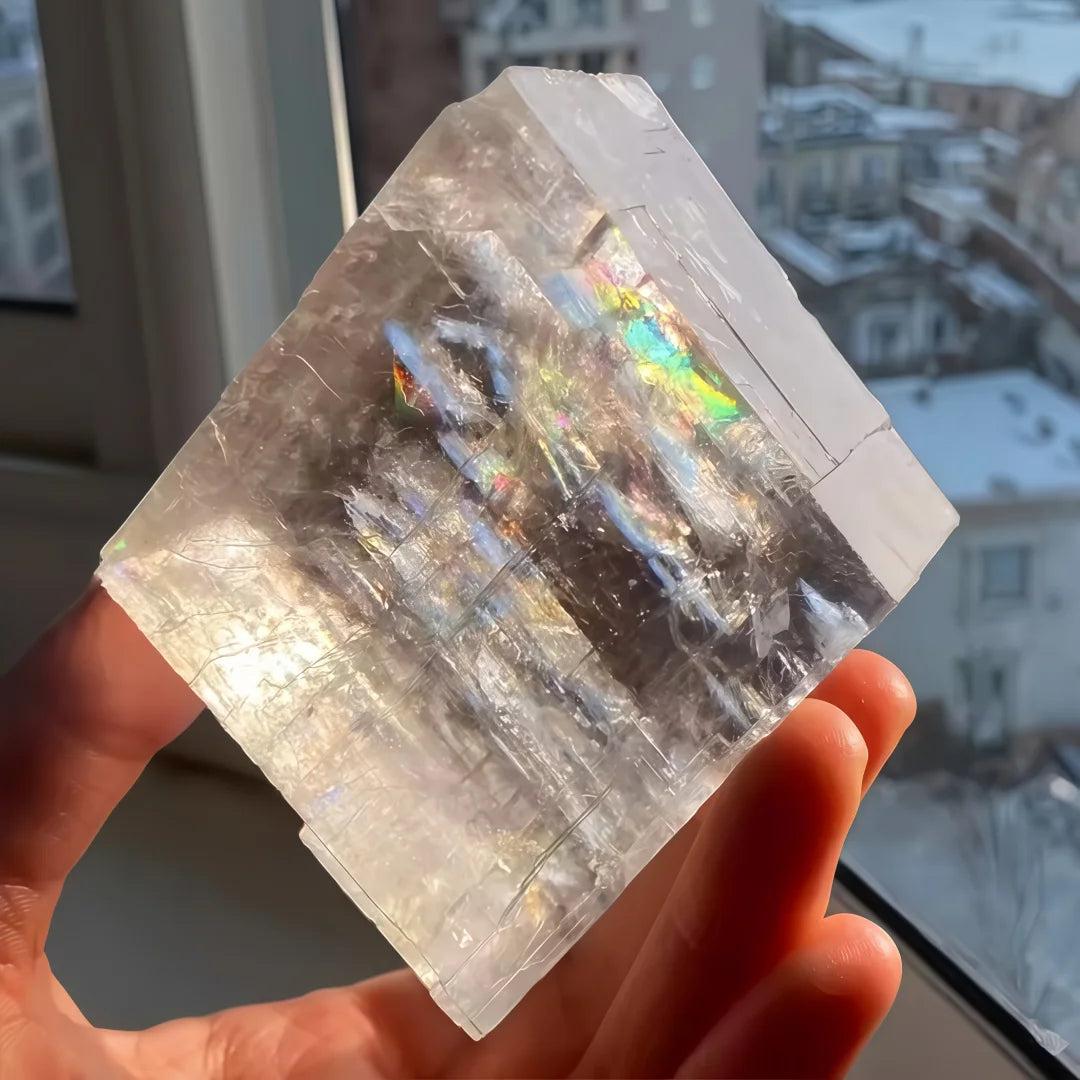 Clear Brazilian optical calcite cube, showing rainbow refractions in sunlight, held in a hand.