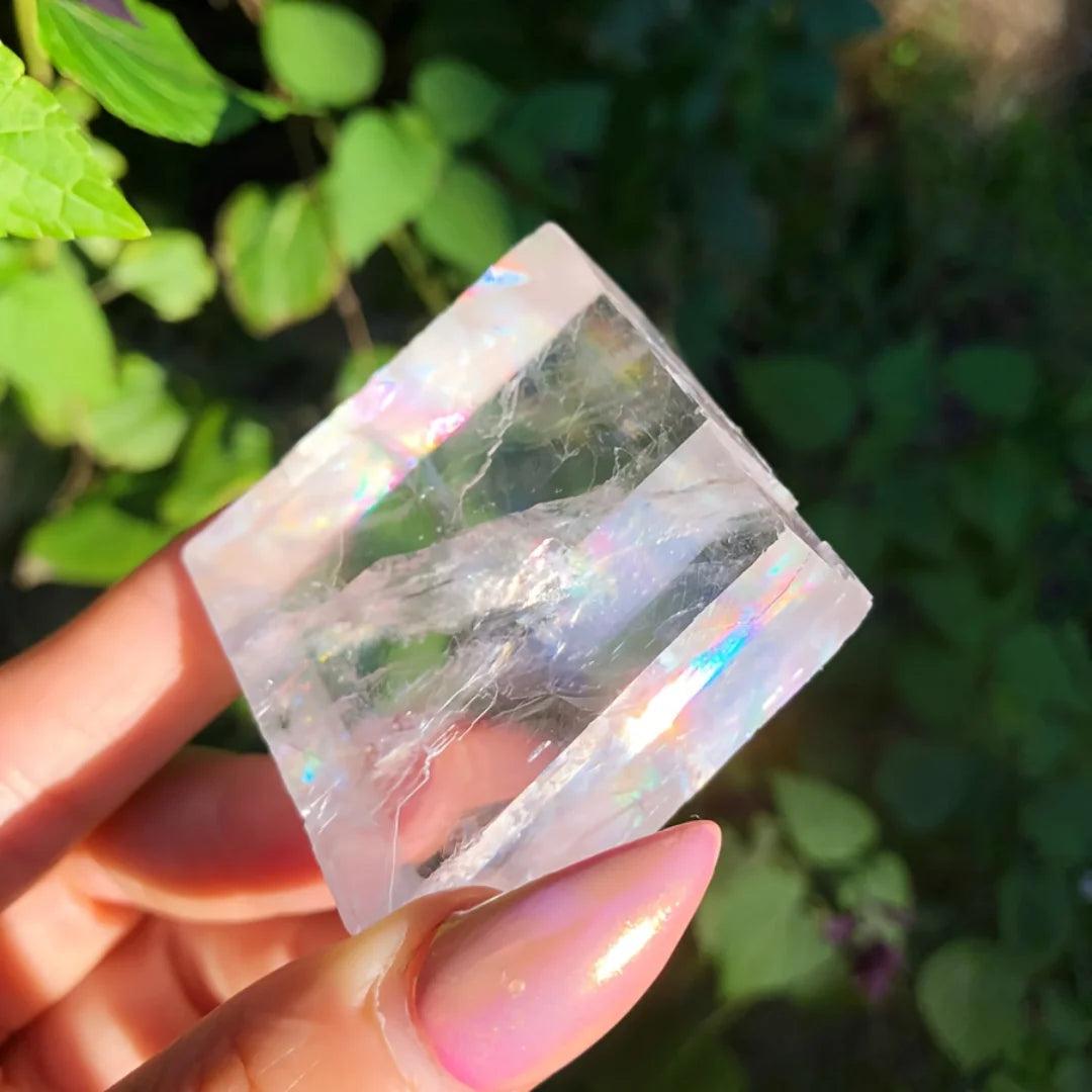 Clear Brazilian Optical Calcite cube with rainbow refractions, held against a green leafy background.