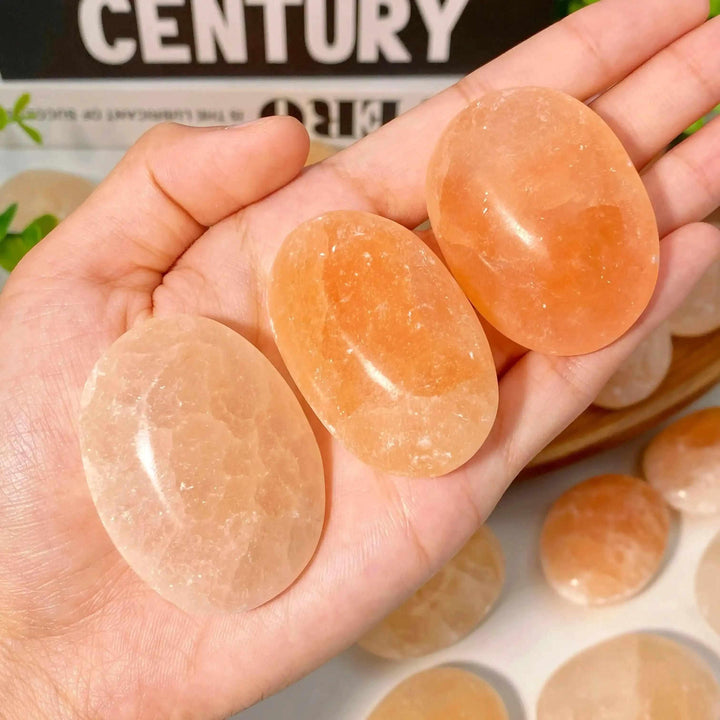 Three smooth, polished Orange Selenite palm stones with a silky peach glow, held in a hand for Sacral Chakra healing.