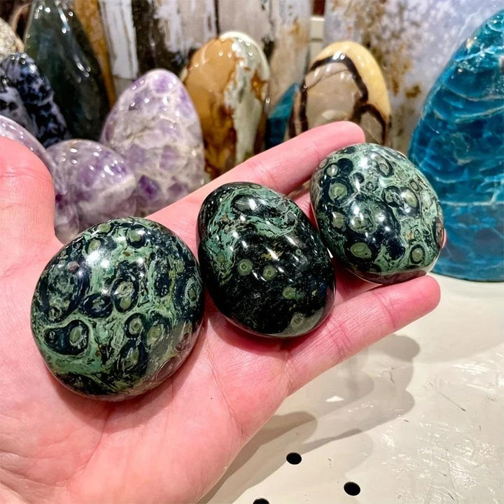 Three polished Peacock Eye crystal palm stones with green and black patterns held in a hand for grounding.