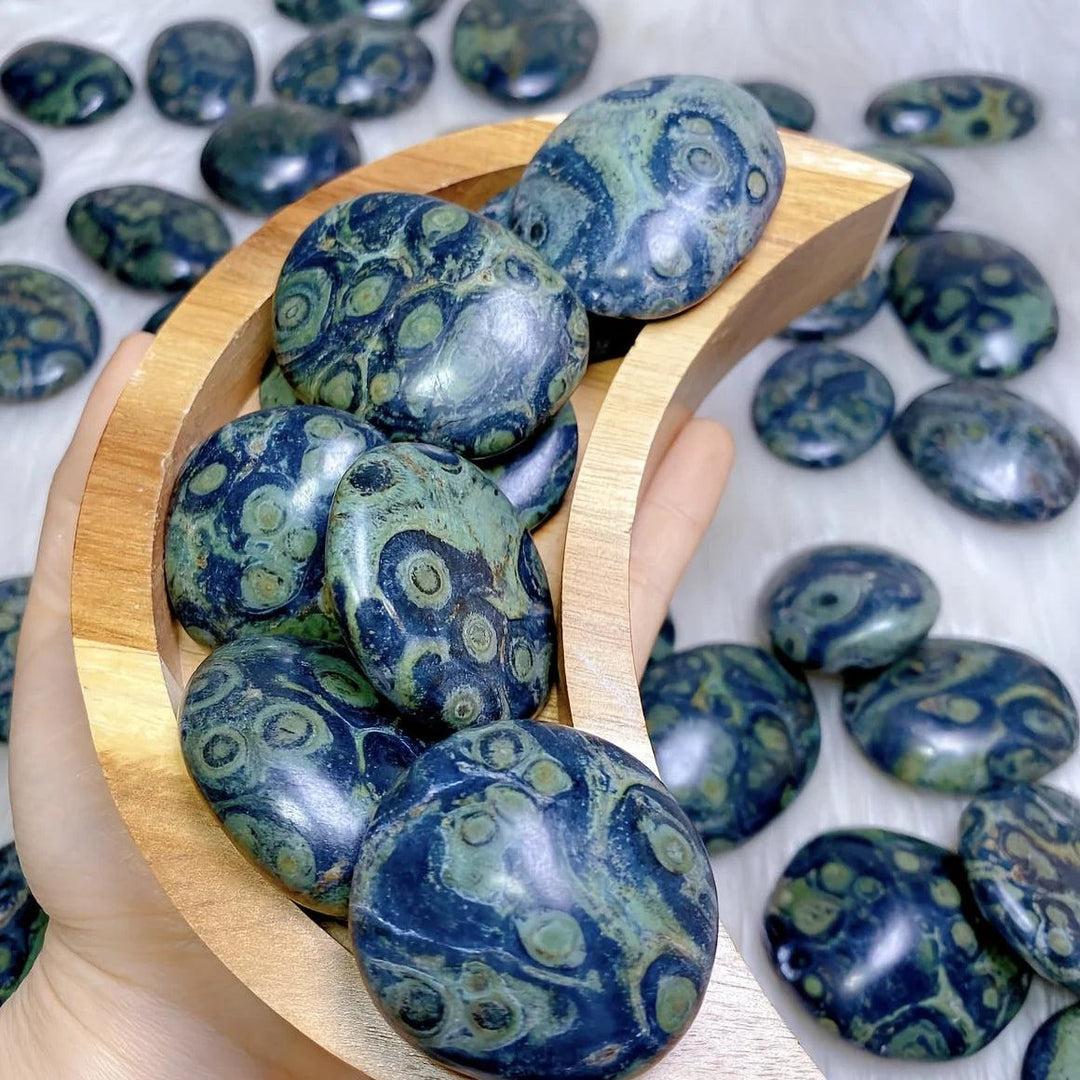 Polished peacock eye crystal palm stones with protective green and blue eye patterns in a wooden moon bowl.