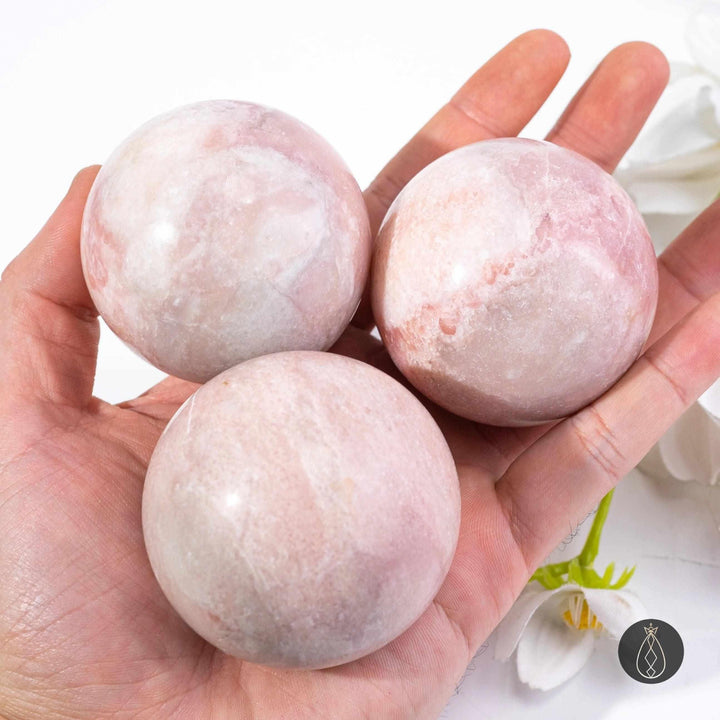 Three polished pink opal spheres in soft blush tones, held in a hand to show their smooth, heart-centered crystal beauty.