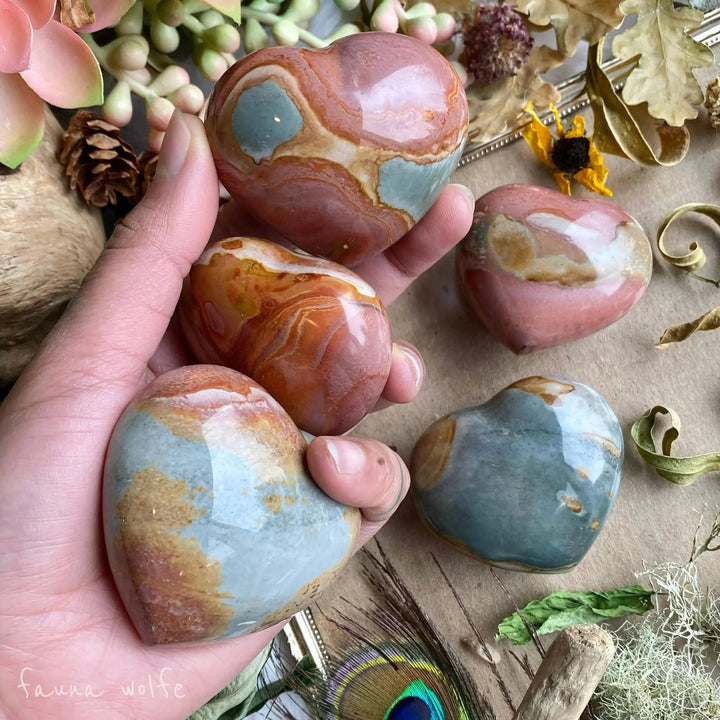 Hand holding five polished Polychrome Jasper hearts with earthy rainbow swirls, promoting passion and grounding.