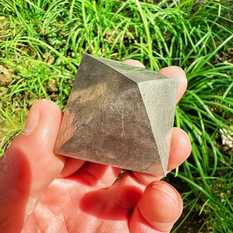 Hand holding a metallic, mirror-bright Pyrite Crystal Pyramid for manifestation and abundance against green grass.