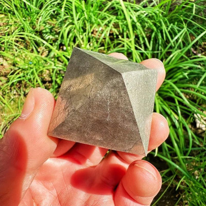 Hand holding a metallic, mirror-bright Pyrite Crystal Pyramid for manifestation and abundance against green grass.