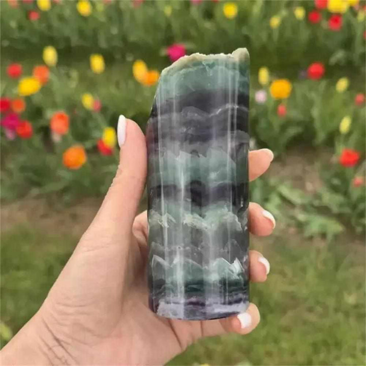 Polished rainbow fluorite crystal lamp with vibrant green and purple banding, held against a colorful floral background.