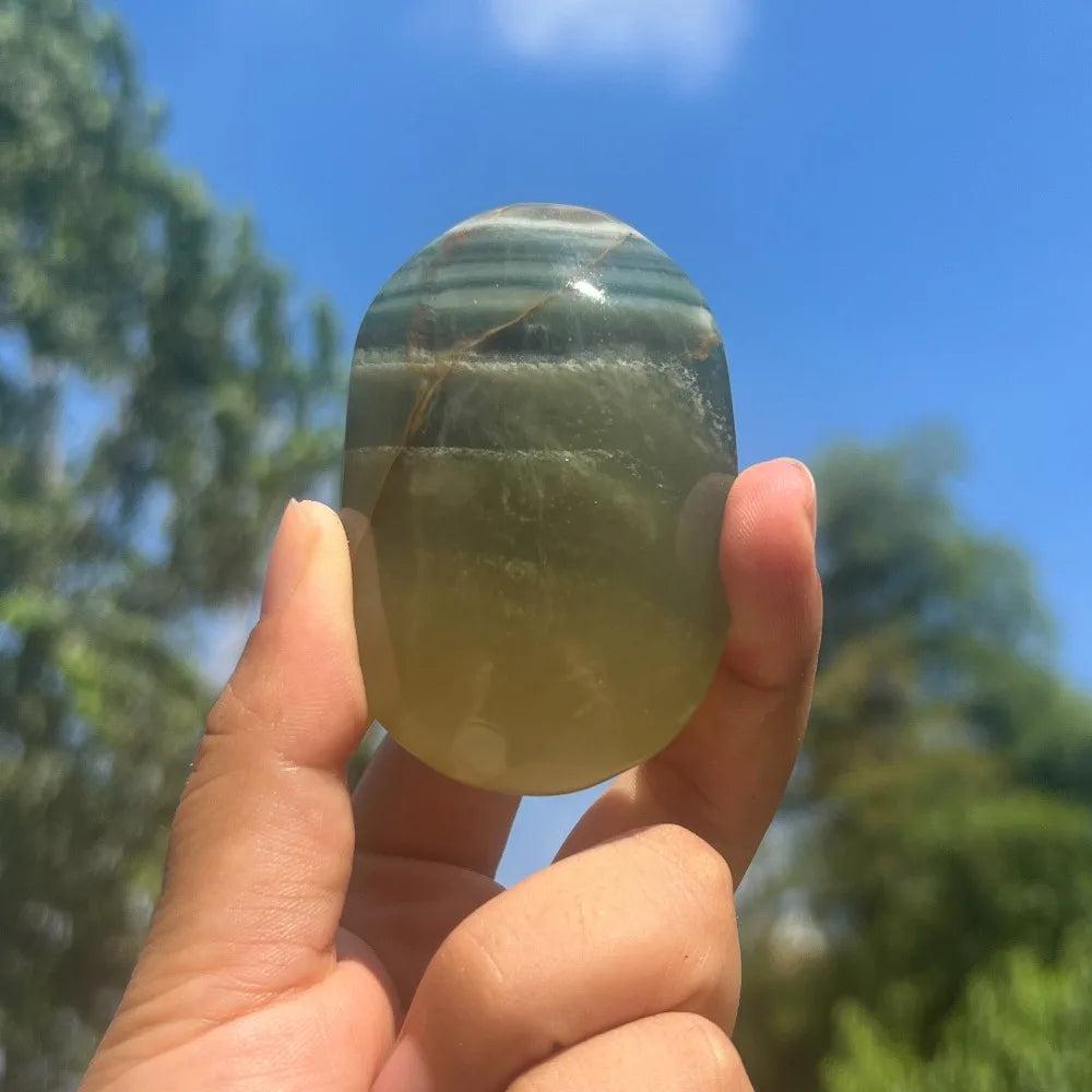 Polished green and blue striped Rainbow Mango Fluorite healing crystal palm stone held against a sunny blue sky.