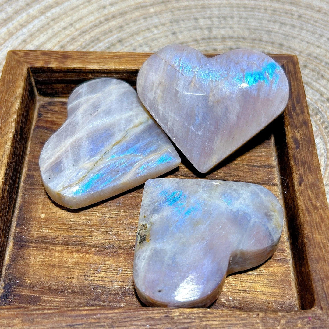 Polished Rainbow Moonstone and Sunstone hearts with milky white tones and iridescent blue flashes in a wooden tray.