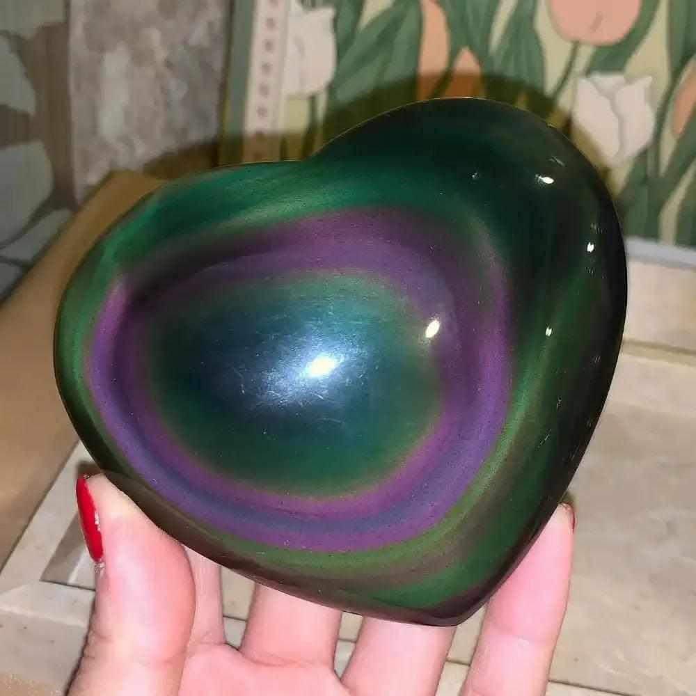 Polished Rainbow Obsidian heart with vibrant violet and green sheen for grounding, held in hand for a smooth, glassy finish.