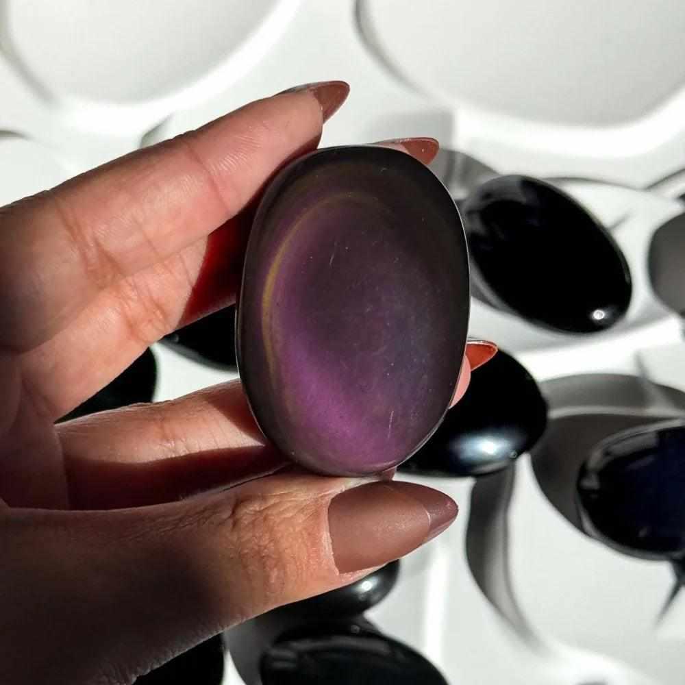 Smooth Rainbow Obsidian palm stone with a glossy black finish and vibrant purple and gold sheen for grounding and protection.