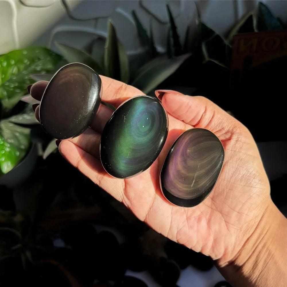 Three smooth Rainbow Obsidian palm stones with glossy black surfaces and vibrant green, purple, and gold sheen bands.