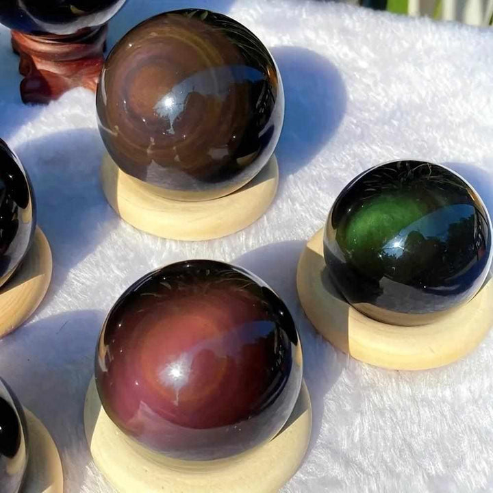 Polished Rainbow Obsidian spheres on wooden stands, showcasing black volcanic glass with shimmering, colorful root chakra bands.