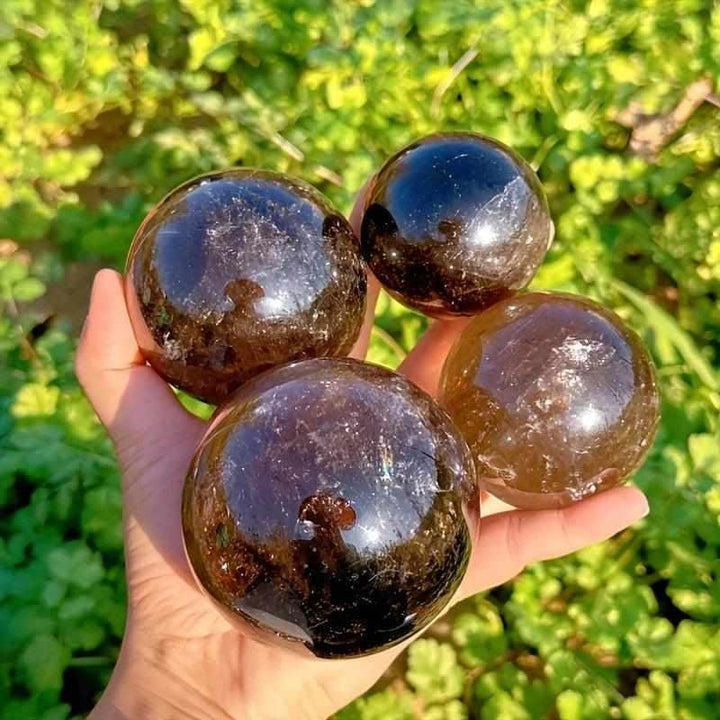 Polished Rainbow Smoky Quartz spheres in warm brown and gray tones, held in hand to showcase grounding energy and rainbows.