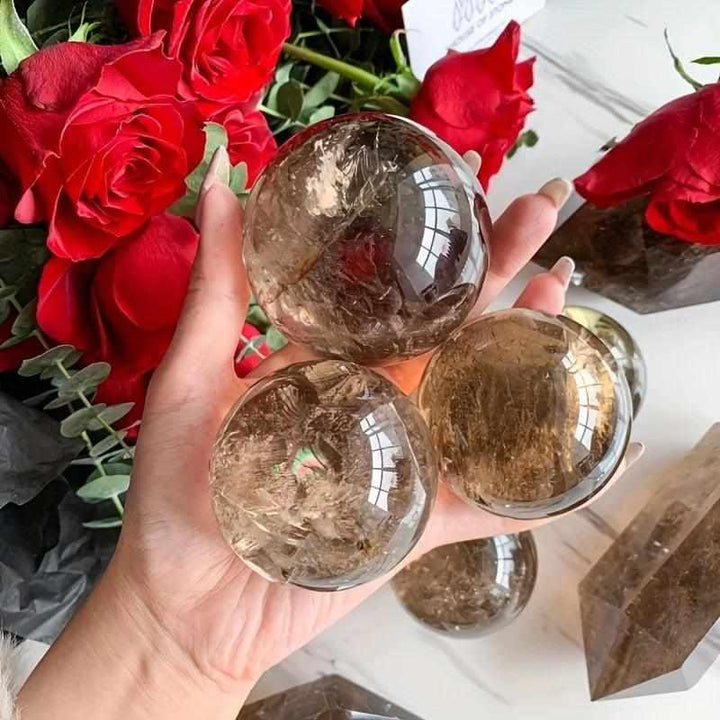 Polished Rainbow Smoky Quartz spheres in warm brown tones, held in hand against red roses for grounding energy.