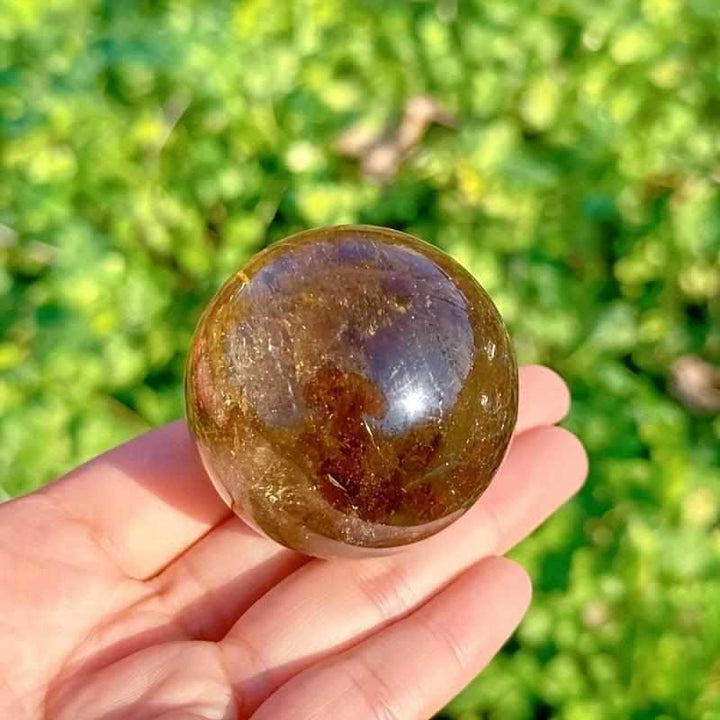 Polished Rainbow Smoky Quartz sphere with warm brown tones and internal flashes held in hand for grounding energy.