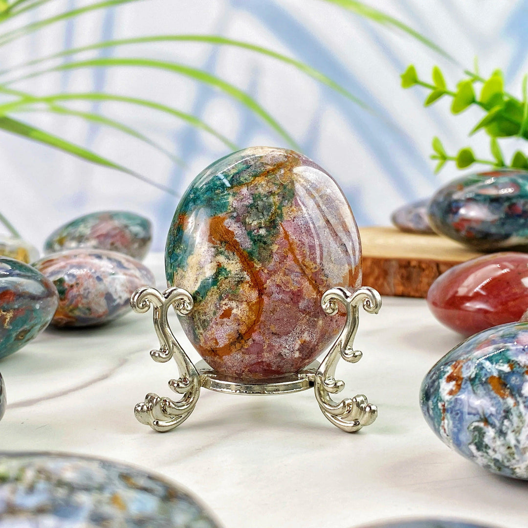 Polished Ocean Jasper palm stone with vibrant green and pink patterns, displayed on an elegant silver stand for decor.