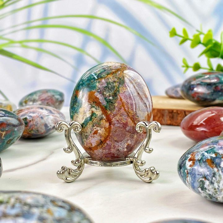 Polished Ocean Jasper palm stone with vibrant green and pink patterns, displayed on an elegant silver stand for decor.