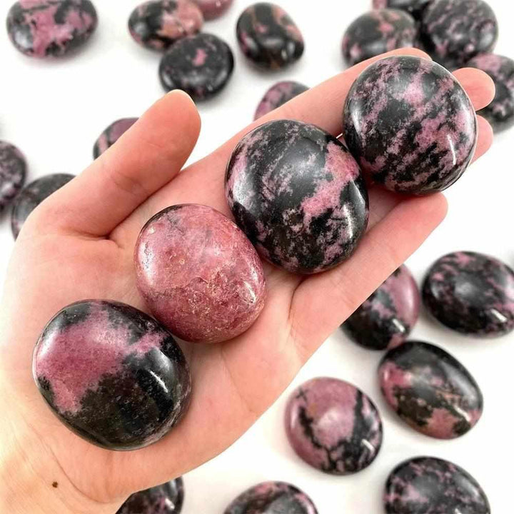 Smooth, polished Rhodonite palm stone with pink hues and black veining, held in hand for heart chakra healing and balance.