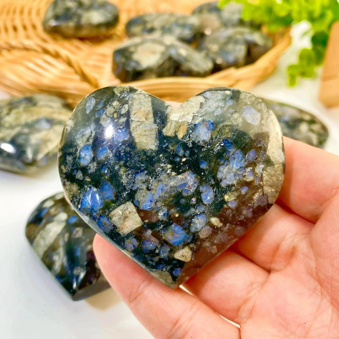 Hand holding a polished blue and black K2 Jasper crystal heart from Crystal Destiny, with more hearts in a basket.