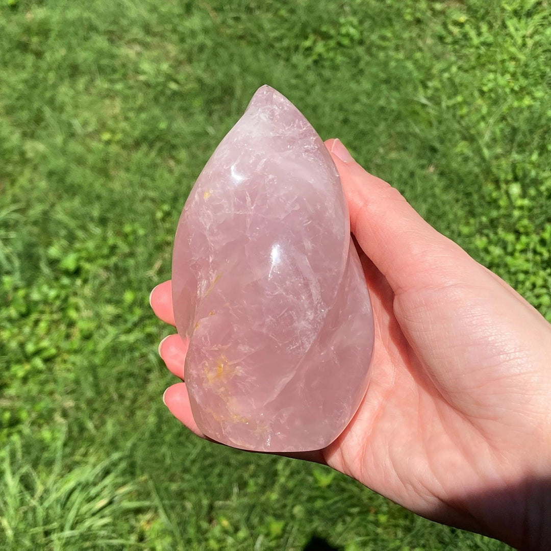 Hand holding a polished, soft pink Rose Quartz crystal torch, tapered upward to a flowing point, against green grass.