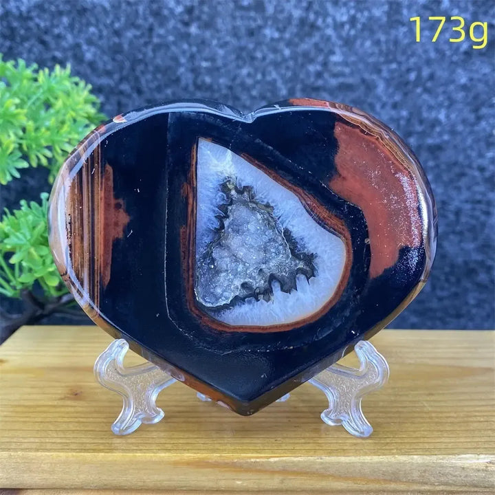 Heart-shaped black and red agate geode with sparkling white crystal cave, 173g, on a stand.