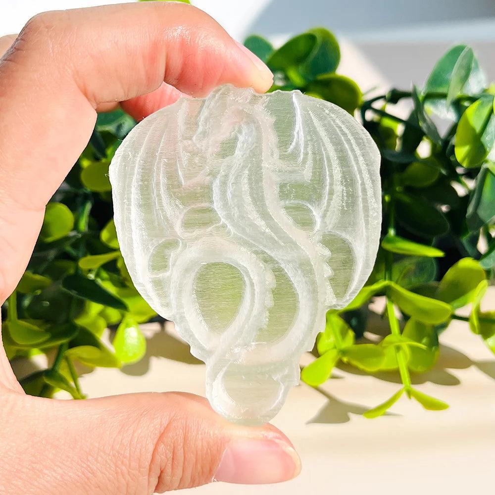 Selenite Crystal Carved Flying Dragon, glowing milky white, held against green foliage.